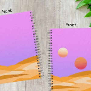 May include: Spiral-bound notebook with a desert landscape design. The cover features a gradient sky in shades of purple and orange, with two suns and sand dunes. The back cover has the same design. The notebook is open, showing the front and back covers.