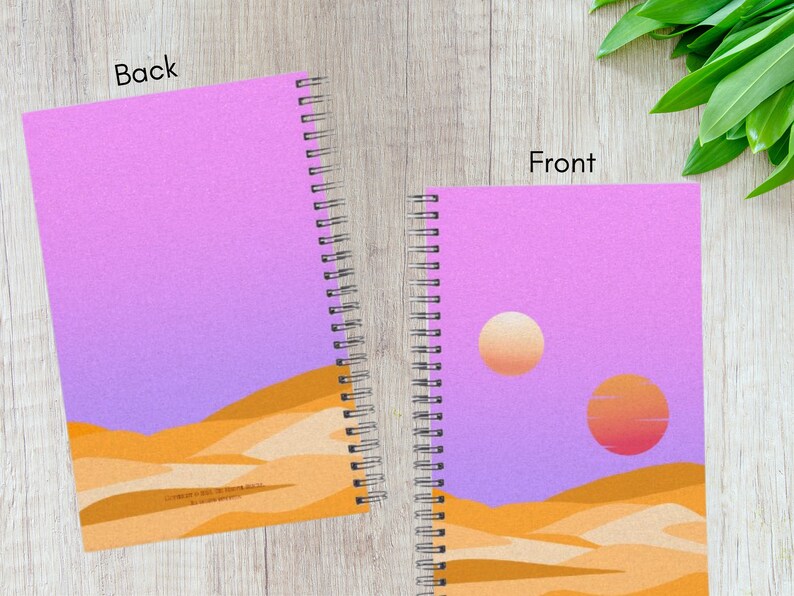 May include: Spiral-bound notebook with a desert landscape design. The cover features a gradient sky in shades of purple and orange, with two suns and sand dunes. The back cover has the same design. The notebook is open, showing the front and back covers.