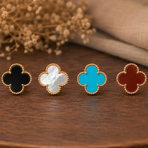 May include: Four clover-shaped stud earrings arranged in a row. Each earring features a gold-toned beaded border surrounding a central inlay. The inlays are black, iridescent white, bright turquoise, and deep reddish-brown, respectively.