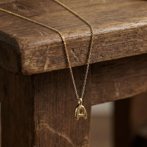 Puede incluir: Un collar dorado con un colgante con la forma de la letra "A". El colgante es de estilo burbuja redondeada. La fina cadena cuelga sobre una superficie de madera.