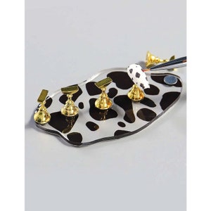 May include: A white and black cow print nail art practice pad with five gold metal nail tips. The pad is made of acrylic and has a smooth surface.