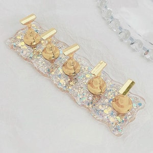 May include: A clear, iridescent nail art display stand with five gold metal nail holders. The stand is decorated with gold and iridescent glitter.