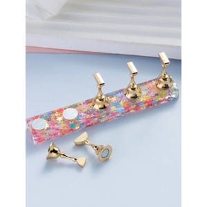 May include: Gold nail art display stand with three nail holders and two extra nail holders. The stand is made of clear resin with colorful glitter and white circles.