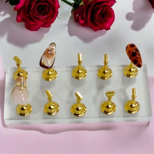 May include: A clear acrylic display stand with eight gold metal nail art display holders. The holders have a round base and a small rectangular top. There are three nail art designs on the display stand: a pink and white nail with a rhinestone, a brown and white nail with a floral design, and a brown and black leopard print nail.