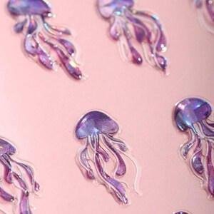 May include: A sheet of iridescent purple and pink holographic jellyfish stickers. The stickers are on a pink background.