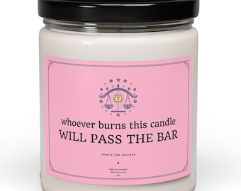 May include: Pink candle with a white label that says "whoever burns this candle WILL PASS THE BAR" and features a Libra zodiac symbol. The label also says "smells like success".