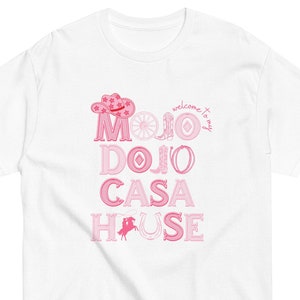 Puede incluir: Camiseta blanca con texto rosa que dice "Welcome to my Mojo Dojo Casa House". El texto está decorado con iconos de temática western como un sombrero de vaquero, una herradura y una bota.