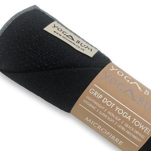 May include: A rolled-up black yoga towel with a grey trim. The towel features a textured surface with grip dots. The label reads "YOGA BUM" and "GRIP DOT YOGA TOWEL". The towel is made of microfiber.