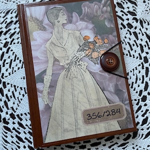 Peut inclure: Un journal relié en cuir marron avec un motif floral représentant une femme en robe tenant un bouquet de fleurs. Le journal a une fermeture à bouton marron et une étiquette avec le numéro 356/284.