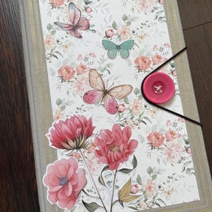May include: A grey journal with a floral design and a pink button closure. The cover features butterflies, flowers, and a floral pattern.