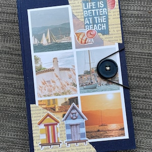 May include: A blue notebook with a collage of images featuring sailboats, houses, and a beach scene. The notebook has a black button closure and a paper bookmark with the text "Life is better at the beach".