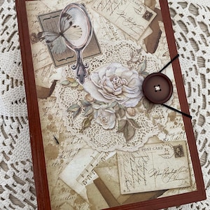 Peut inclure: Un journal décoratif avec une couverture marron, orné d'éphémères vintage. La couverture présente un papillon, un miroir, une rose, des cartes postales et une fermeture à gros bouton. Le design incorpore une nappe en dentelle et des éléments de papier vieilli.