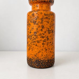 Puede incluir: Un jarrón cilíndrico de cerámica con un color base naranja vibrante y un esmalte texturizado con motas de color marrón oscuro. El jarrón presenta un borde superior grueso y redondeado, y una base con un esmalte de textura rugosa marrón oscuro.