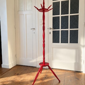 Op de afbeelding: Een hoge, felrode houten kapstok met een centrale gedraaide paal, een driepotige voet en een rond bovengedeelte met meerdere haken voor het ophangen van kleding. De structuur is volledig afgewerkt met een egale rode lak.