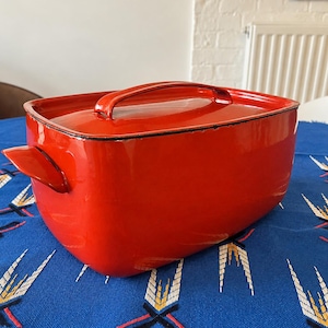 Pode incluir: Uma assadeira retangular de metal esmaltado em vermelho brilhante com tampa a condizer. O recipiente possui duas pegas laterais e uma pega central na tampa, com um rebordo preto fino a delinear as extremidades da base.