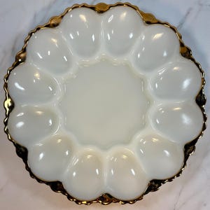 May include: A white, scalloped egg serving dish with a gold-colored trim. The dish has 12 egg-shaped indentations around the edge and a circular center. The dish is made of a glossy material.