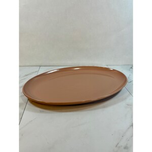May include: A smooth, oval-shaped serving platter with a solid, muted terracotta-colored finish. The dish features a wide, shallow basin and a slightly raised rim, presenting a clean, uniform appearance without any patterns or decorative textures.