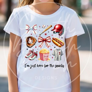 Op de afbeelding: Wit T-shirt met honkbalthema-afbeeldingen, waaronder een honkbal, knuppel, helm, handschoen, schoenplaatjes, popcorn en een hotdog. Het shirt heeft de tekst "I'm just here for the snacks" en is versierd met strikken en sterren.