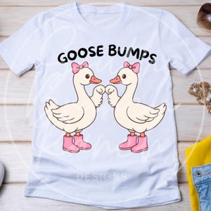 Può includere: T-shirt bianca con un design a cartoni animati di due oche con fiocchi e stivali rosa, che si tengono per mano. Il testo "GOOSE BUMPS" è stampato sopra le oche. La maglietta ha uno scollo tondo.