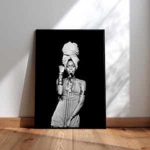May include: A black and white framed print of a singer performing on stage. The singer is wearing a head wrap, a dress, and jewellery. She is holding a microphone and has her fist raised. The print is leaning against a white wall on a wooden floor.