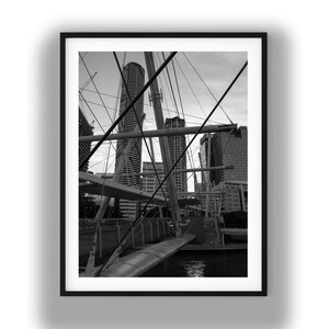 May include: Black and white photograph of a modern pedestrian bridge with a cityscape backdrop. The bridge features a unique cable-stayed design, with tall buildings and a cloudy sky in the background. The image is framed in a black border.