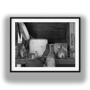 Könnte beinhalten: Schwarz-Weiß-Fotografie einer Sammlung von Vintage-Kunstutensilien. Ein großer, weißer Farbeimer steht neben verschiedenen Gläsern, Flaschen und Schachteln. Das Bild ist gerahmt und hat eine rustikale, gealterte Ästhetik.