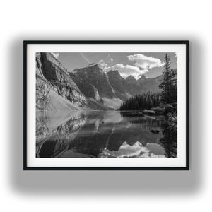 May include: A black and white framed landscape photograph featuring a calm lake reflecting jagged mountain peaks and a dense forest of pine trees under a cloudy sky. The composition is centered within a simple black frame with a white mat border.