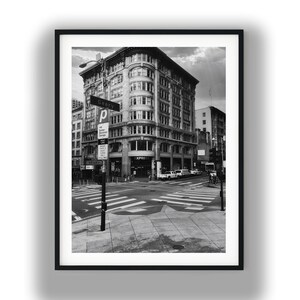 May include: Black and white framed photograph of a city intersection with a large building. A street sign reads "Geary". The crosswalk is visible, and there are cars and people in the distance. The sky is overcast.
