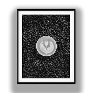 Op de afbeelding: Zwart-wit ingelijste print met een latte met een hartvormig schuimontwerp, geplaatst op een bed van donker gebrande koffiebonen. De opname van bovenaf benadrukt het contrast tussen de lichte koffie en de donkere bonen.