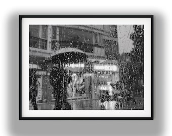 May include: A black and white framed wall art print depicting a rainy city street scene viewed through a window pane covered in raindrops. The composition features a silhouette of a person holding an umbrella, blurred storefront lights, and building architecture in the background, all captured in a monochromatic, moody aesthetic.