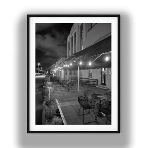 Può includere: Fotografia in bianco e nero di un ristorante all'aperto di notte. L'immagine mostra tavoli e sedie disposti sotto una tenda con luci a corda. Un edificio è sullo sfondo.