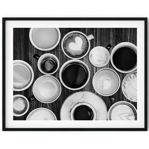 May include: A black and white photograph of a selection of coffee cups and mugs on a wooden table. The cups are filled with coffee, cappuccino, and latte. Some of the cups have a heart design in the foam.
