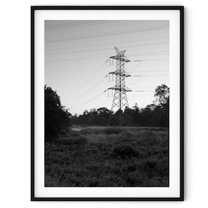 Op de afbeelding: Een zwart-witfoto van een hoge elektriciteitsmast in een veld. De mast is gemaakt van metaal en heeft veel draden die er vanaf lopen. Het veld is bedekt met gras en er zijn bomen op de achtergrond.