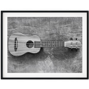 May include: A black and white image of a ukulele with a wooden body and fretboard. The ukulele is resting on a textured wooden surface. The instrument has four strings and tuning pegs.