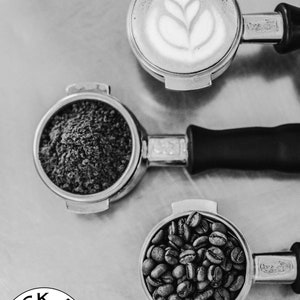 K&ouml;nnte beinhalten: Drei Espressoportafilter aus Metall, einer gef&uuml;llt mit Kaffeepulver, einer mit ganzen Kaffeebohnen und einer mit einem Latte-Art-Design.