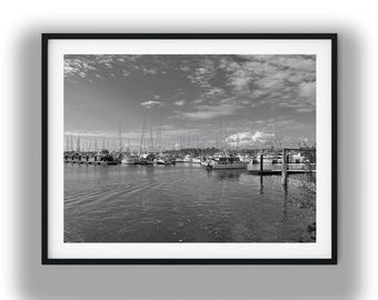 May include: Black and white photograph of a marina filled with sailboats and yachts. The boats are docked in the water, with masts reaching towards a cloudy sky. The image is framed in a black border.