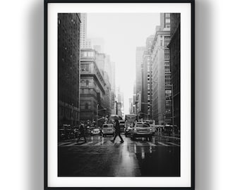 May include: A black and white framed photograph of a city street scene during a rainy day. A person with an umbrella walks across a wet crosswalk surrounded by tall buildings and cars. The composition captures a busy urban intersection in monochrome.