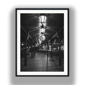May include: A black and white framed wall art print depicting a train station platform at night. The scene features a long covered walkway with rows of dark pillars, overhead hanging lanterns, and a central round clock suspended from the ceiling.