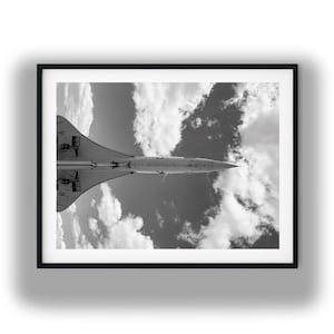 May include: A black and white framed photograph showing the underside of a supersonic passenger jet in flight. The aircraft is positioned horizontally against a sky filled with scattered clouds, highlighting its pointed nose and delta wings.