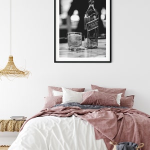 May include: A black and white framed wall art print featuring a bottle of Johnnie Walker Black Label scotch whisky and a glass with ice cubes. The artwork is displayed in a simple black frame hanging on a plain white wall above a bed.