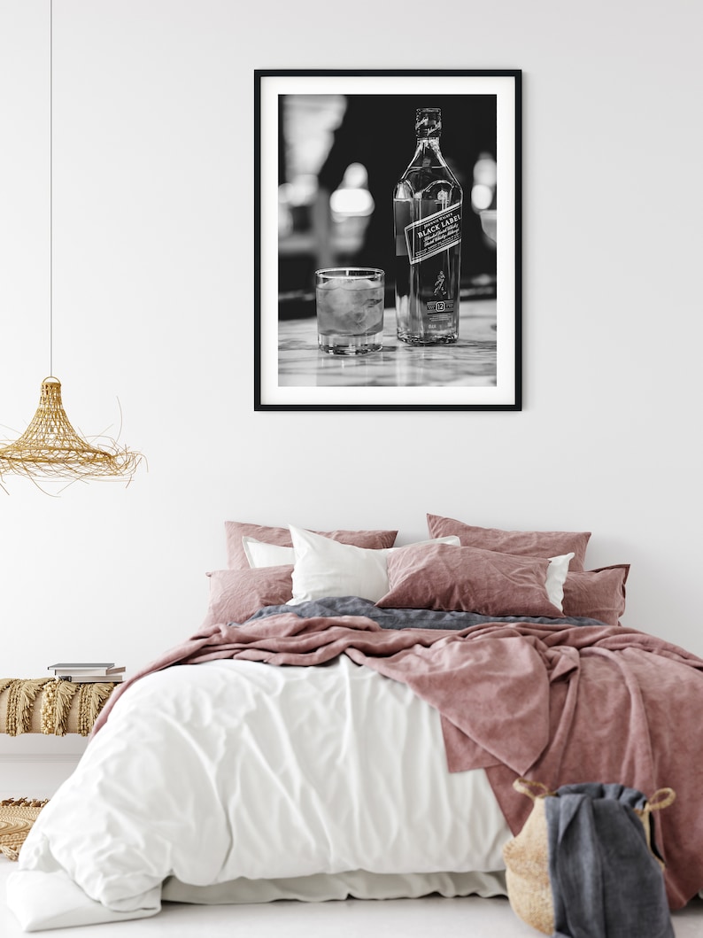 May include: A black and white framed wall art print featuring a bottle of Johnnie Walker Black Label scotch whisky and a glass with ice cubes. The artwork is displayed in a simple black frame hanging on a plain white wall above a bed.