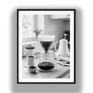 Puede incluir: Una fotografía en blanco y negro de un gotero de café de vidrio sobre una jarra de vidrio con el número 02 impreso en el frente. También se ven una pequeña cuchara de cerámica con café molido, una jarra pequeña y una tetera de cuello de cisne.
