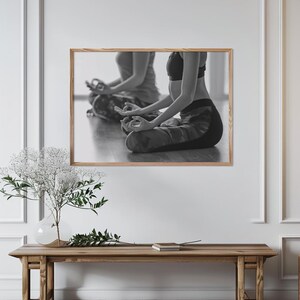 Könnte beinhalten: Ein gerahmter Schwarz-Weiß-Wandkunstdruck, der zwei Personen in einer sitzenden Yoga-Pose mit Händen in Mudra-Haltung zeigt. Das Kunstwerk ist in einem hellen Holzrahmen gerahmt und an einer weißen Wand über einem Holzkonsolentisch montiert.