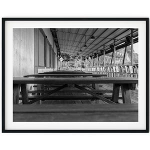 Op de afbeelding: Een zwart-witfoto van een rij lege houten picknicktafels onder een overdekt terras. De tafels zijn in een rij geplaatst, waarbij het perspectief van de foto ze in de verte doet verdwijnen. Het terras is gemaakt van houten planken en heeft een metalen dak.