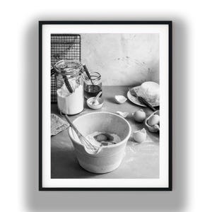 May include: A black and white photograph of a baking scene featuring a large mixing bowl with a wire whisk, a glass jar filled with flour, a small jar of liquid, a plate with a round pastry, and several cracked eggshells on a flat surface.