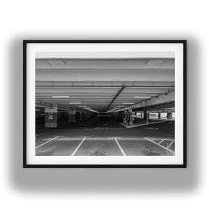 May include: Black and white photograph of an empty parking garage. The image is framed with a black border. The garage has multiple levels, with rows of parking spaces marked by white lines. Overhead, there are fluorescent lights.