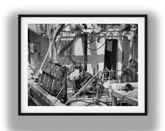 May include: A black and white photograph of a fishing boat deck featuring complex rigging, heavy-duty machinery, coiled ropes, and metal structures. The scene captures the weathered texture of the vessel with various mechanical components and cables.
