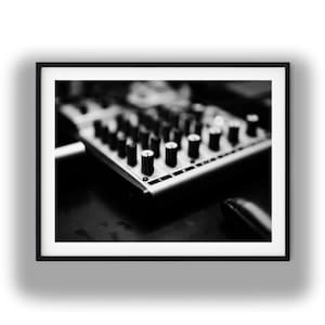 May include: Black and white image of a sound mixer with numerous knobs and sliders. The mixer is rectangular with a silver finish and is set against a dark background. The knobs are black and cylindrical.