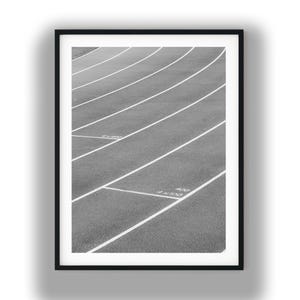 May include: A black and white framed print showing a textured running track with curved white lane markings. The surface features white painted text including 4x400 and 4x100 markings on the asphalt. The composition is captured from a high angle.