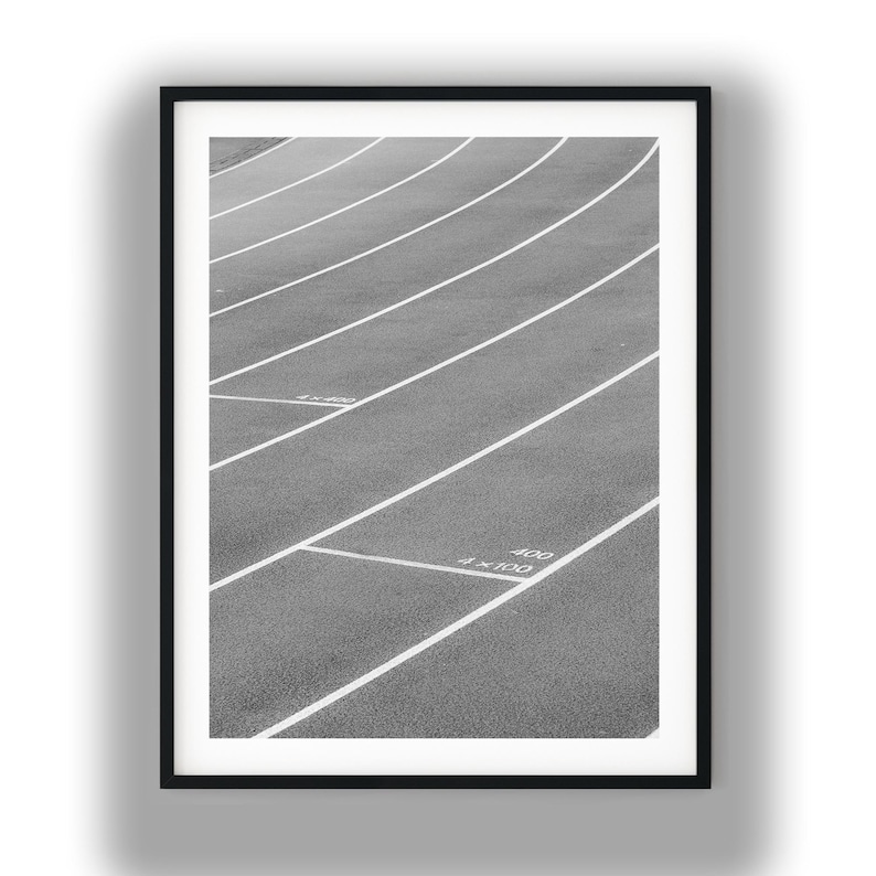 May include: A black and white framed print showing a textured running track with curved white lane markings. The surface features white painted text including 4x400 and 4x100 markings on the asphalt. The composition is captured from a high angle.